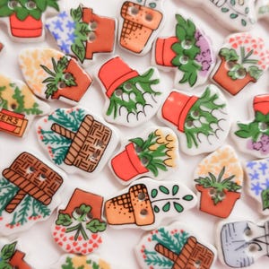 Pode incluir: Uma coleção de botões de cerâmica com tema de jardim. Os botões apresentam várias plantas em vasos, flores e cestos em tons de laranja, verde e branco. Alguns botões têm a palavra "FLOWERS".