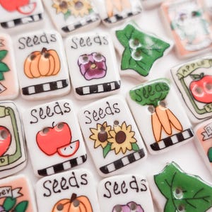 May include: A collection of decorative ceramic buttons with various garden-themed designs. Each button features a hand-painted illustration of vegetables, flowers, or fruits, with the word "Seeds" and other text. The buttons are white with black and white checkered borders.