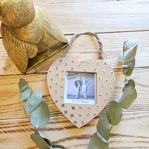 May include: A pink heart-shaped picture frame with a 3x3 inch photo opening. The frame is decorated with gold glitter and yellow and black bee stickers. The text "PolkaDots Designs" and "3" x 3" Photo" is printed on the frame.