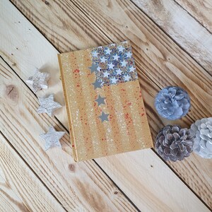 May include: A yellow notebook with a red, white, and blue American flag design. The notebook is on a wooden surface with white stars and pine cones.
