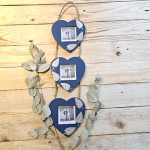 May include: A navy blue, heart-shaped photo frame with three frames suspended from a rope. Each frame holds a small black and white photo and is decorated with silver leaf accents. The frames are displayed against a wooden background.