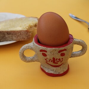May include: A whimsical, handmade egg cup with a smiling face design, holding a brown egg. The cup has two handles and a red rim. A slice of buttered toast and two spoons are also visible on a yellow surface.