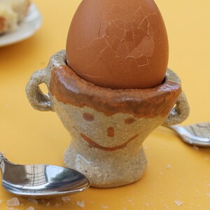 May include: A boiled brown egg in a ceramic egg cup with a smiling face design. The egg cup has a brown rim and handles. Two spoons and a plate of bread are on a yellow surface.