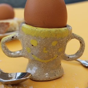 May include: A handmade ceramic egg cup with a smiling face design, holding a brown egg. The cup has a yellow rim and handles. Two spoons and a plate of bread are on a yellow surface.