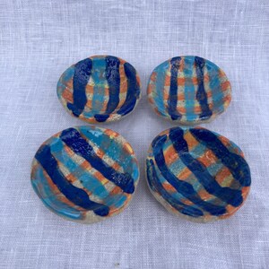 May include: Four small, hand-painted ceramic dishes. Each dish is a light beige color with a blue, orange, and turquoise striped pattern. The dishes are arranged in a square formation, showcasing their unique, artisan design.