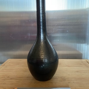 Puede incluir: Un jarrón de cerámica negro con un cuello largo y delgado y una base redondeada. El jarrón tiene una superficie texturizada y un acabado ligeramente iridiscente.
