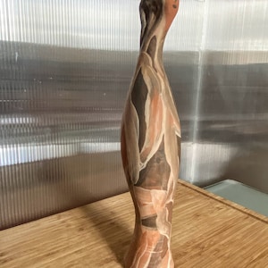 Pode incluir: Uma escultura de cerâmica de uma figura humana estilizada com um design pintado em marrom, branco e rosa. A figura tem um pescoço longo e fino e uma cabeça redonda com um rosto simples.
