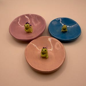 Op de afbeelding: Drie kleine keramische schalen in roze, blauw en perzik. Elke schaal heeft een klein geel beeldje met paarse accenten in het midden. De schalen zijn rond met een licht verhoogde rand.