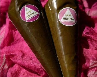 May include: Two henna cones, filled with a dark brown paste, are displayed on a pink fabric. Each cone has a pink and white label that reads "gopi HENNA". One label says "Darkness Blend" and the other says "Original Henna Blend".