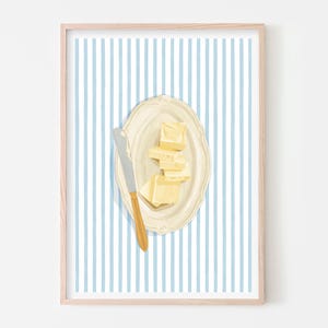 May include: A framed art print featuring a still life of butter on a plate with a butter knife. The butter is cut into cubes and sits on a cream-colored plate. The background has vertical blue and white stripes.