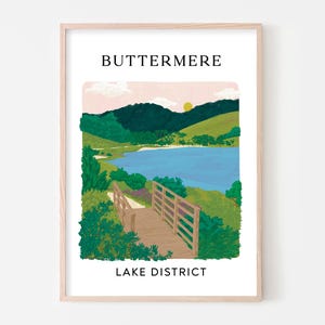 Può includere: Una stampa artistica incorniciata con un'illustrazione paesaggistica di Buttermere nel Lake District. L'immagine mostra una passerella in legno in primo piano, un lago blu, colline verdi e un cielo rosa pallido con un sole giallo. Il testo recita BUTTERMERE LAKE DISTRICT.