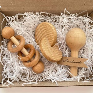 May include: A set of three wooden baby toys in a box with white shredded paper. The toys include a rattle, a teether, and a ring. The box has a brown paper label that says "Handmade".