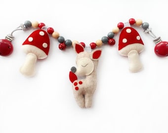 Pode incluir: Um móbile de brinquedo para bebê de feltro com um veado, dois cogumelos vermelhos e contas de madeira. O móbile tem dois clipes de plástico vermelho em cada extremidade.
