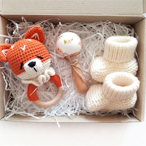Puede incluir: Una caja de regalo llena de artículos para bebés, que incluyen un sonajero de zorro de crochet, un sonajero de madera con una parte superior de cerámica blanca y un par de botines tejidos.