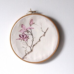 May include: A round, off-white fabric art piece featuring a hand-painted floral design. The artwork depicts pink magnolia flowers and branches, framed by a natural wood embroidery hoop. The piece is mounted on a white wall.