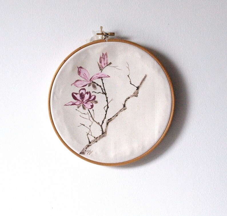 May include: A round, off-white fabric art piece featuring a hand-painted floral design. The artwork depicts pink magnolia flowers and branches, framed by a natural wood embroidery hoop. The piece is mounted on a white wall.