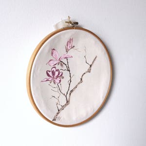 May include: A round, wooden embroidery hoop holds a canvas with a watercolor painting of pink magnolia blossoms and branches. The artwork is attached with a cream-colored ribbon.