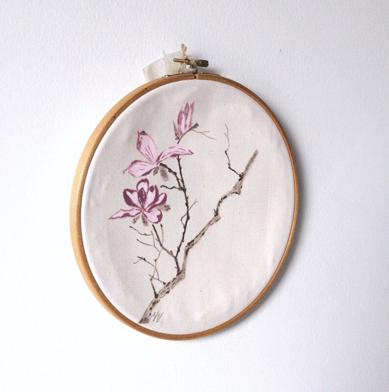 May include: A round, wooden embroidery hoop holds a canvas with a watercolor painting of pink magnolia blossoms and branches. The artwork is attached with a cream-colored ribbon.