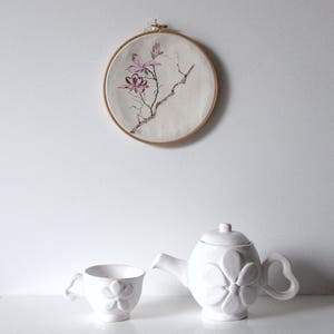 May include: A white ceramic teapot and teacup set with a floral design. The teapot has a rounded shape with a flower-shaped design on the front and a heart-shaped handle. The teacup has a matching floral design. Above, a round embroidery hoop displays a floral painting.