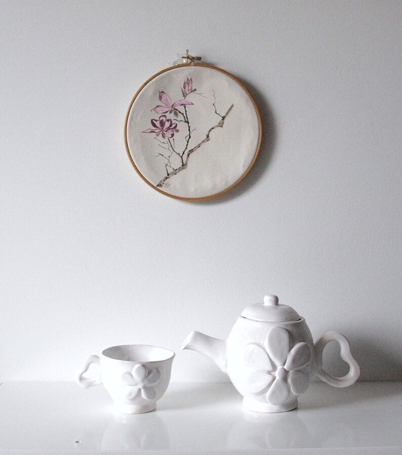 May include: A white ceramic teapot and teacup set with a floral design. The teapot has a rounded shape with a flower-shaped design on the front and a heart-shaped handle. The teacup has a matching floral design. Above, a round embroidery hoop displays a floral painting.