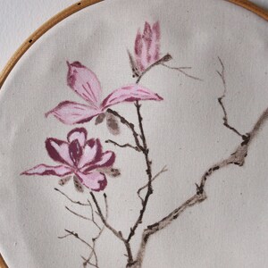 May include: Hand-painted floral artwork on canvas, stretched within a wooden embroidery hoop. The design features pink magnolia blossoms and branches against a neutral background. The artwork is a decorative piece for home decor.