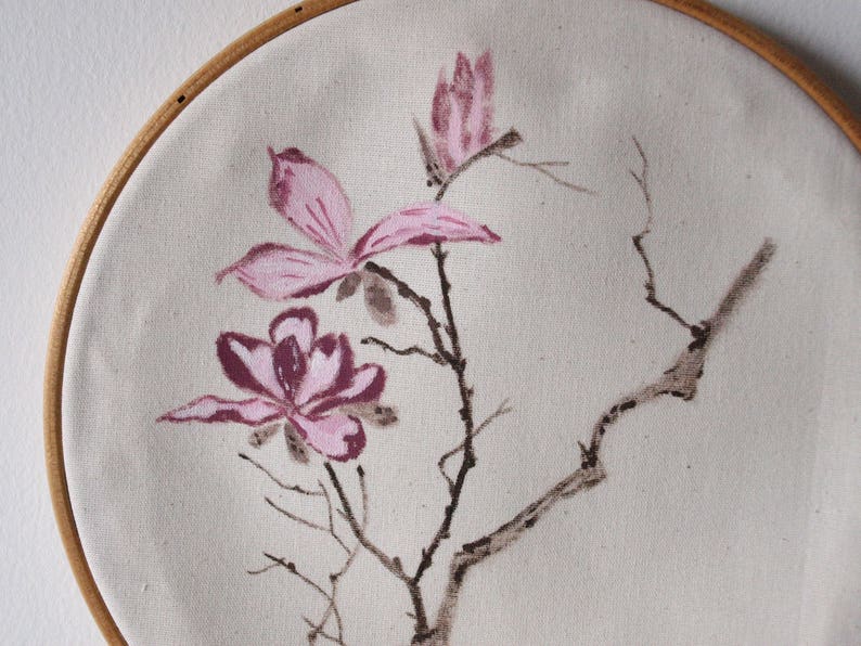 May include: Hand-painted floral artwork on canvas, stretched within a wooden embroidery hoop. The design features pink magnolia blossoms and branches against a neutral background. The artwork is a decorative piece for home decor.