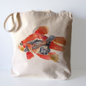 May include: A beige canvas tote bag featuring a watercolor painting of a goldfish. The fish is primarily orange, red, and gray, with detailed fins and scales. The bag has a simple design with long handles for carrying.