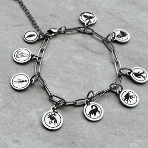 Puede incluir: Una pulsera de plata con dijes circulares, cada uno con una ilustración negra. Los dijes muestran varios símbolos, incluyendo animales y figuras, unidos por una cadena. La pulsera tiene un cierre de mosquetón.