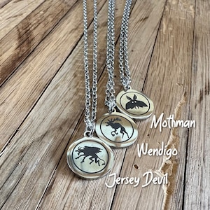 May include: Three silver necklaces with circular pendants. Each pendant displays a black silhouette of a cryptid: Mothman, Wendigo, and Jersey Devil. The pendants are on a wooden surface, with the cryptid names in white script.
