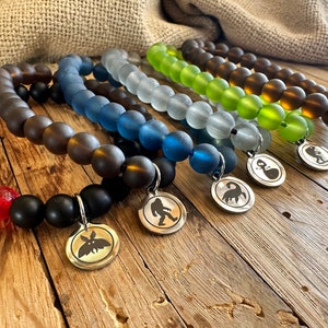 May include: A collection of beaded bracelets in various colors, including black, brown, blue, green, and clear. Each bracelet features a unique silver charm with a silhouette design. The bracelets are displayed on a rustic wooden surface.