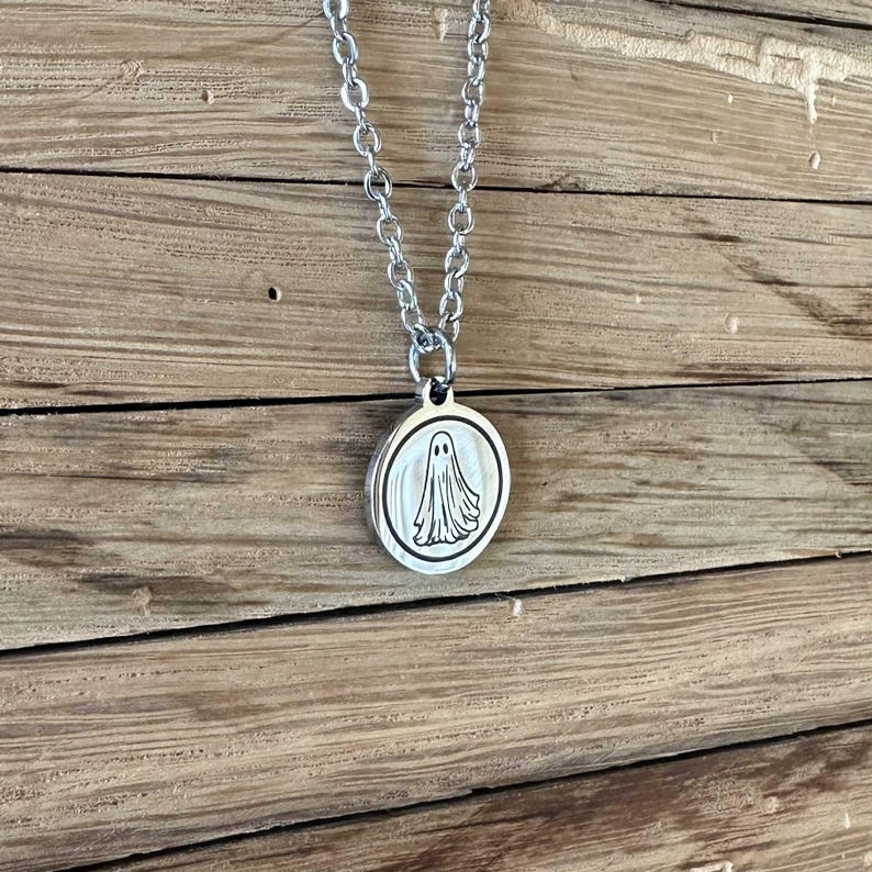May include: A silver-coloured metal necklace featuring a circular pendant with a black engraved ghost illustration. The pendant hangs from a matching silver-coloured link chain, showcasing a simple design with a polished finish on the round charm.