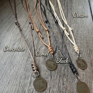 May include: Four necklaces with circular pendants featuring an engraved animal design, each attached to a cord in chocolate, camel, black, or cream. Each necklace includes a branded tag labelled The Crow's Court and adjustable sliding knots.