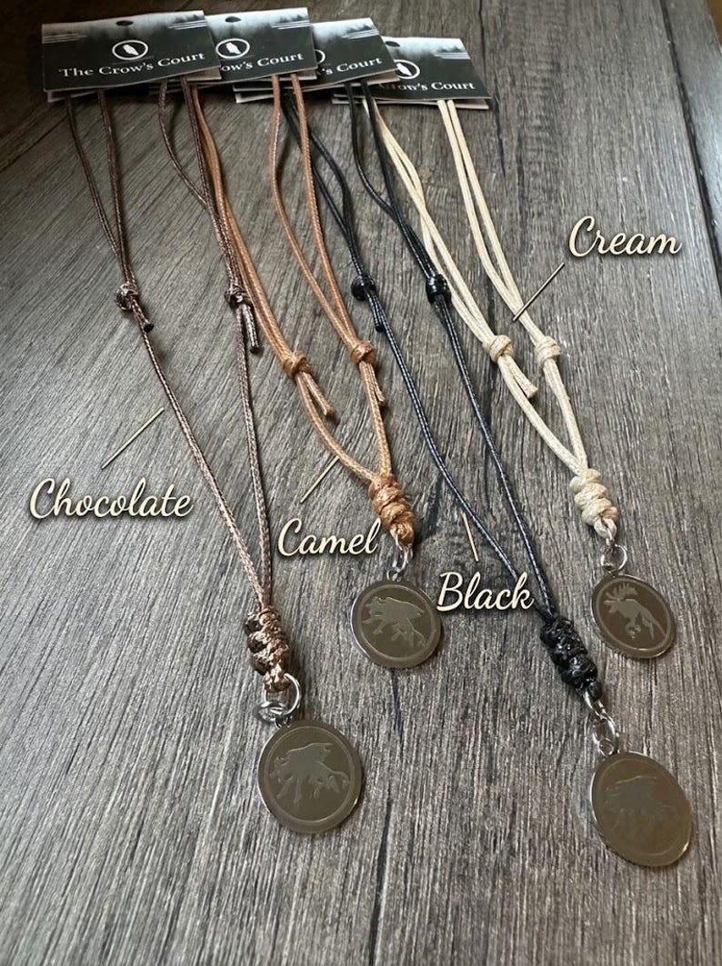 May include: Four necklaces with circular pendants featuring an engraved animal design, each attached to a cord in chocolate, camel, black, or cream. Each necklace includes a branded tag labelled The Crow's Court and adjustable sliding knots.
