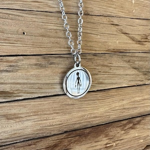 May include: A silver-toned metal pendant necklace featuring a circular charm with a polished finish. The centre of the charm displays a black silhouette graphic of an extraterrestrial figure. The pendant hangs from a matching silver-toned chain.