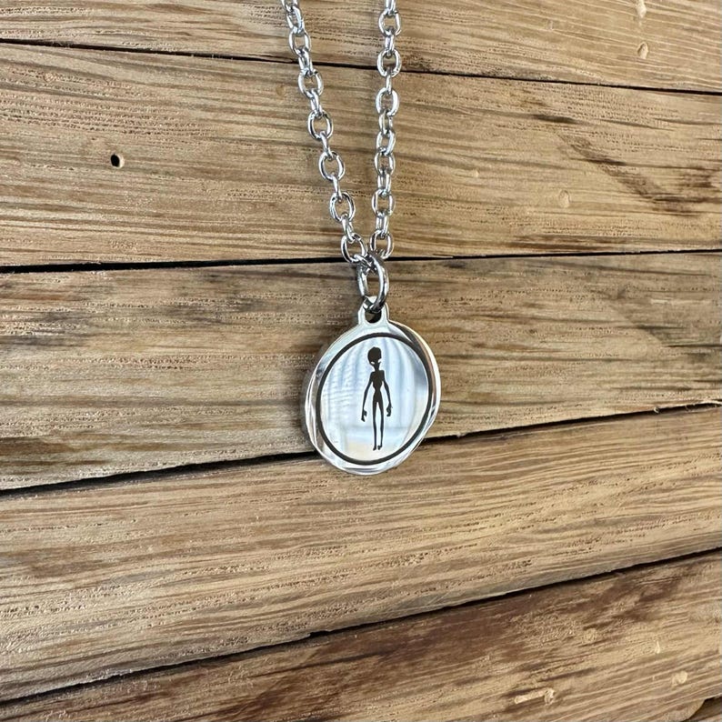 May include: A silver-toned metal pendant necklace featuring a circular charm with a polished finish. The centre of the charm displays a black silhouette graphic of an extraterrestrial figure. The pendant hangs from a matching silver-toned chain.