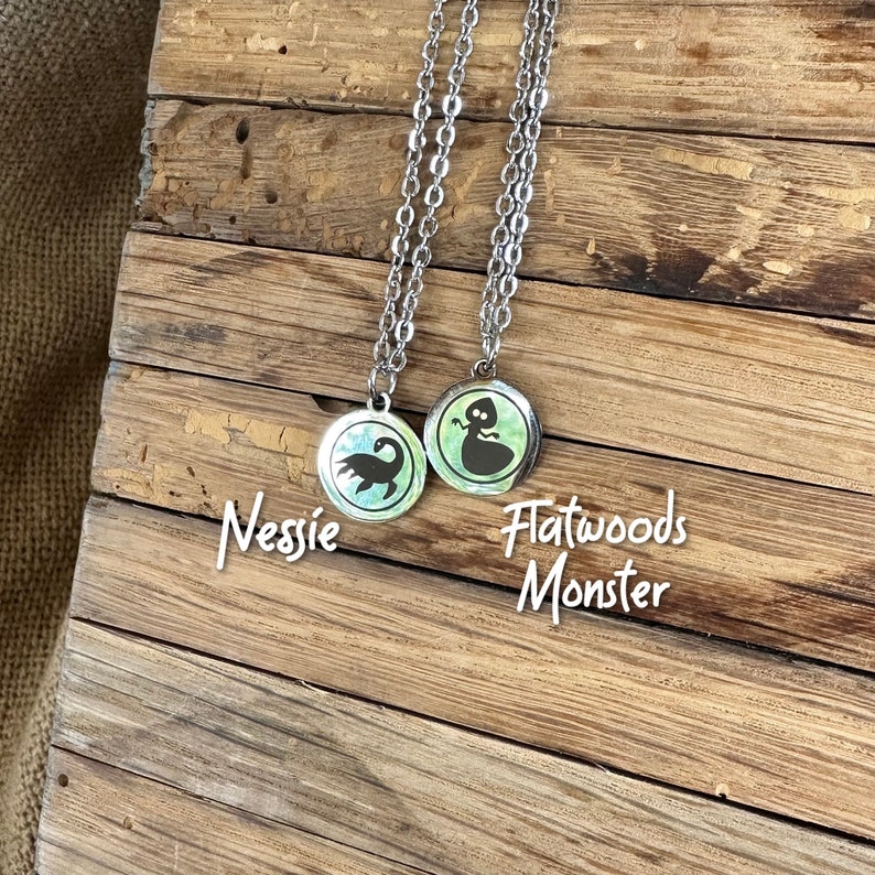 May include: Two silver-toned necklaces featuring circular pendants with green marbled backgrounds. One pendant displays a black silhouette of Nessie, and the other shows a black silhouette of the Flatwoods Monster, with the text Nessie and Flatwoods Monster.