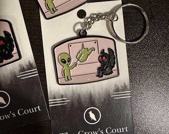 May include: A metal keyring featuring a light pink enamel charm with a green alien pointing at a map of West Virginia while a black Mothman figure sits nearby. The charm is attached to a silver split ring and mounted on a branded display card.