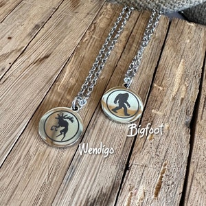 May include: Two silver-toned circular pendant necklaces with matching link chains. One pendant features a black silhouette of a Wendigo, and the other features a black silhouette of Bigfoot, with the text Wendigo and Bigfoot below.