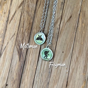 May include: Two silver-toned necklaces featuring circular pendants with a light green background. One pendant displays a black silhouette of Mothman, and the other displays a black silhouette of Frogman, with the text Mothman and Frogman.