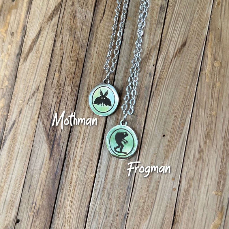 May include: Two silver-toned necklaces featuring circular pendants with a light green background. One pendant displays a black silhouette of Mothman, and the other displays a black silhouette of Frogman, with the text Mothman and Frogman.