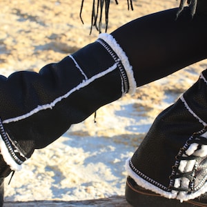 May include: Black leather leg warmers with white faux fur trim and stitching. The leg warmers are worn over black tights.