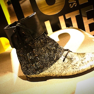 May include: A black and gold ankle boot with a lace-up closure and a decorative floral pattern. The boot has a chunky heel and a rounded toe.