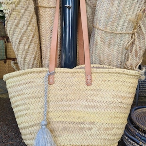 May include: A large, woven straw tote bag with leather handles and a silver tassel. The bag is made of natural straw and has a herringbone pattern.