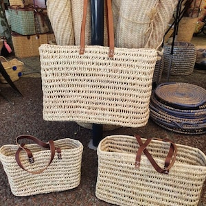 Könnte beinhalten: Drei geflochtene Strohtaschen mit Ledergriffen. Die Taschen sind in verschiedenen Größen und Formen. Die größte Tasche ist eine rechteckige Tasche mit einer breiten Basis. Die mittlere Tasche ist eine rechteckige Tasche mit einer schmaleren Basis. Die kleinste Tasche ist eine rechteckige Tasche mit einer schmalen Basis.