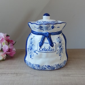 Pode incluir: Um pote de cerâmica branco com um desenho floral azul e branco. O pote tem a forma de uma bolsa e tem uma fita azul amarrada em volta da parte superior. O pote tem a palavra "Cebollas" escrita nele.