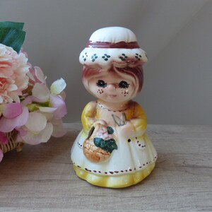 Può includere: Una statuetta in ceramica di una ragazza che indossa un cappello bianco e marrone, un vestito giallo con pois bianchi e tiene un cestino di fiori.