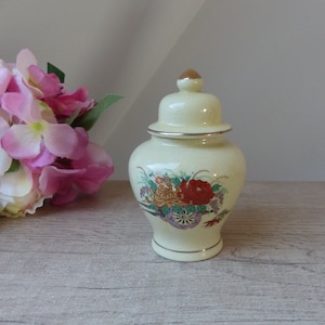 Peut inclure: Un petit pot en céramique de couleur crème avec un motif floral. Le pot a un couvercle et est décoré de fleurs rouges, orange, jaunes et violettes. Le pot est posé sur une surface en bois brun clair.