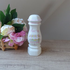 Peut inclure: Un distributeur de sucre en céramique blanche avec un motif floral rose et jaune et le mot "Sucre" écrit sur le devant. Le distributeur est posé sur une surface en bois.