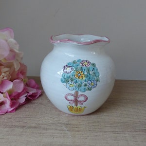 Könnte beinhalten: Eine weiße Keramikvase mit rosafarbenem Rand und handbemaltem Blumendesign. Die Vase zeigt einen blauen Baum mit rosa und gelben Blumen und einer rosa Schleife.