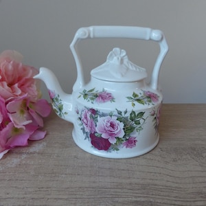 Può includere: Teiera in ceramica bianca con un design floreale con rose rosa e rosse. La teiera ha un coperchio con un pomo decorativo e un manico.